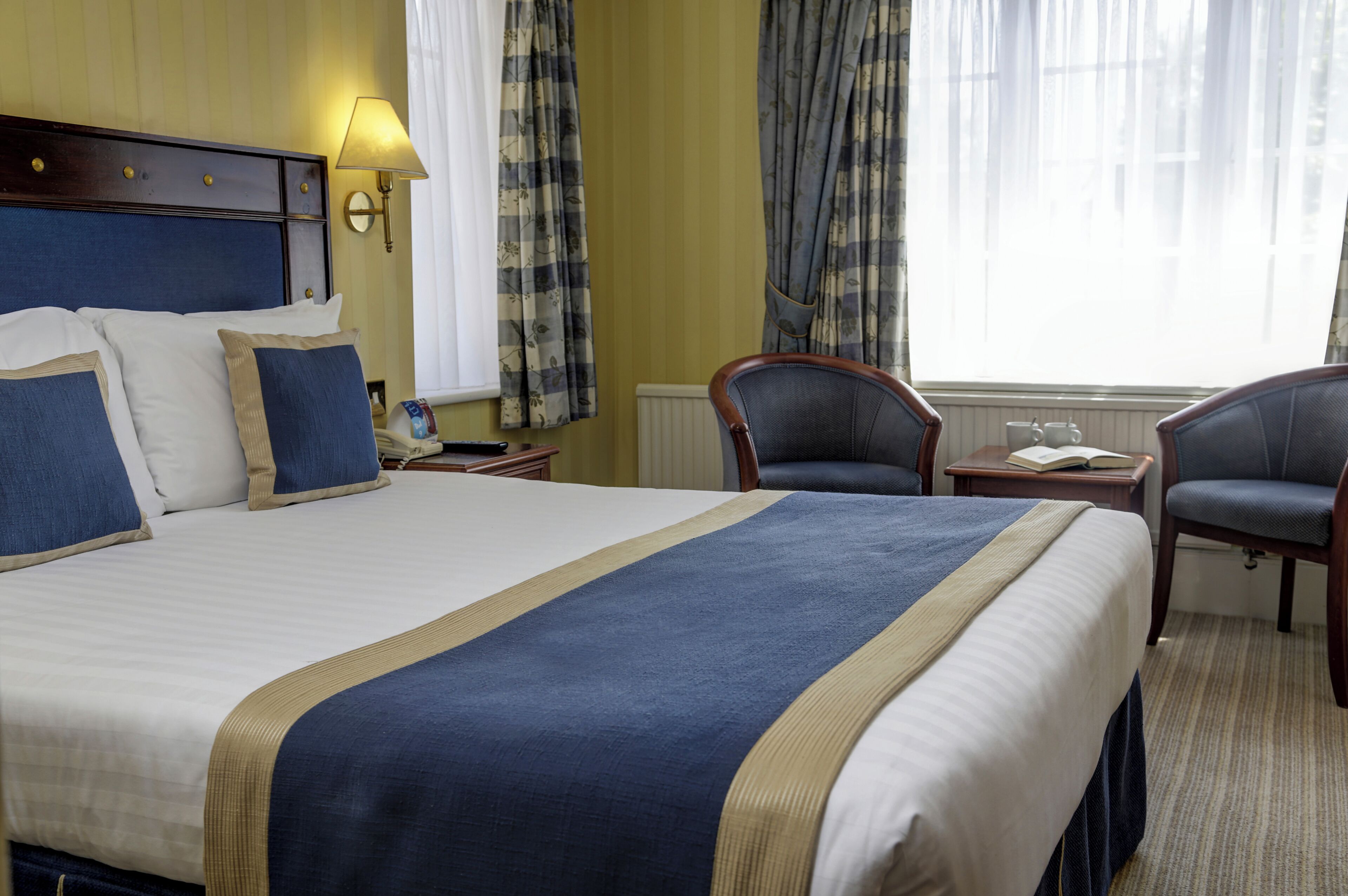 A hotel room with a large bed, two chairs, a nightstand, a lamp, and a window with curtains.