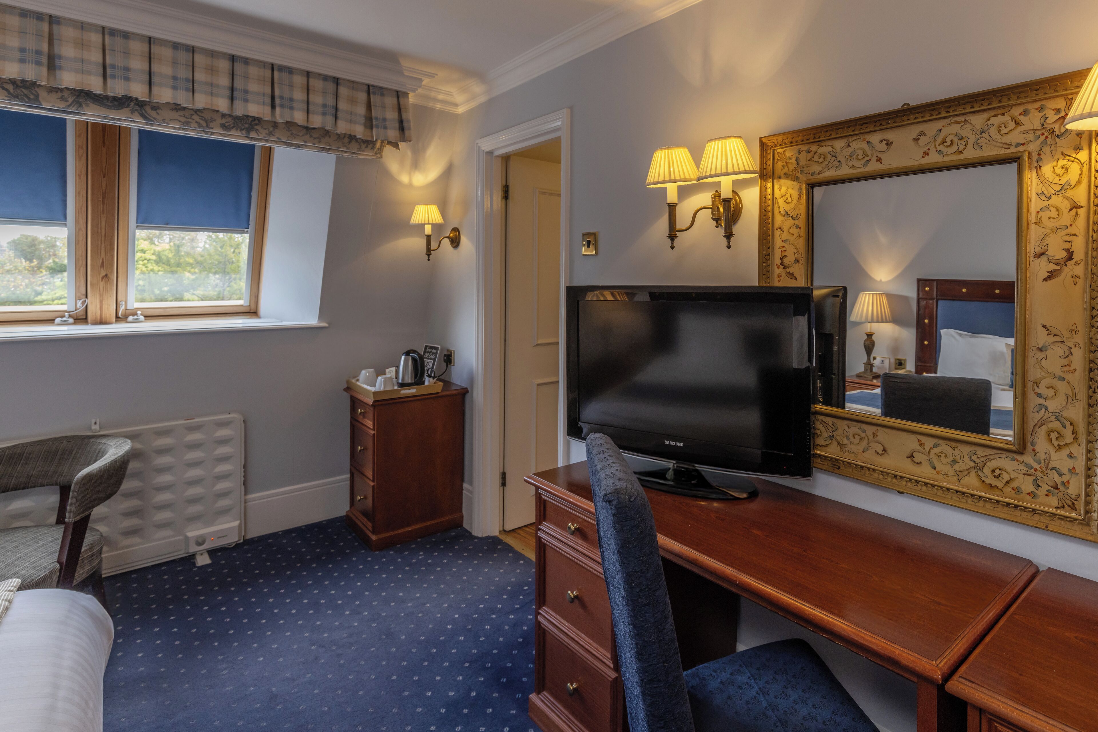 A hotel room with a bed, a desk with a television, a dresser, and a window with blinds.