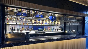 Una zona de bar con estantes llenos de botellas de licores y vasos, una barra y una pantalla digital.