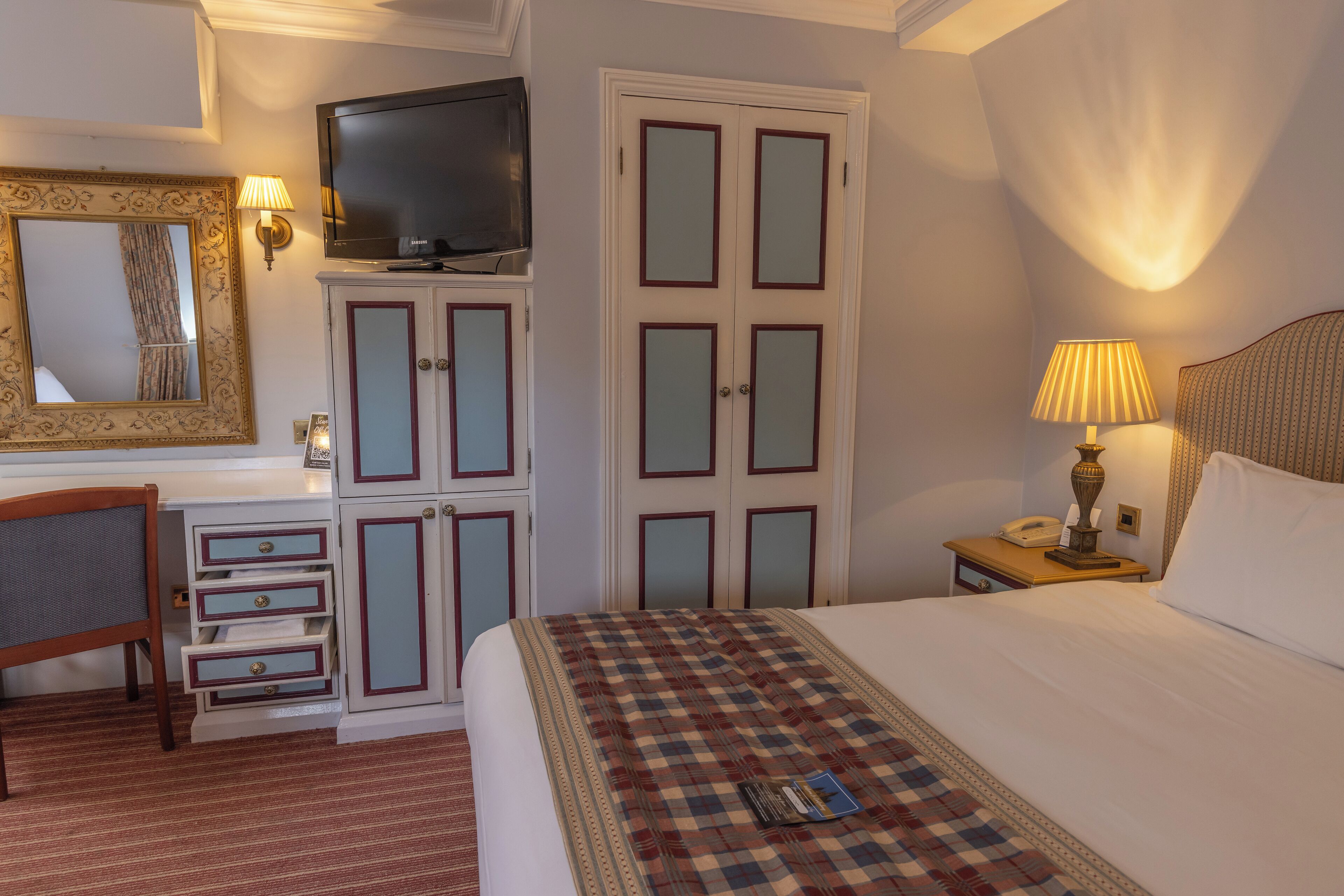 A hotel room with a bed, a desk with drawers, a television, a wardrobe, and a bedside table with a lamp.