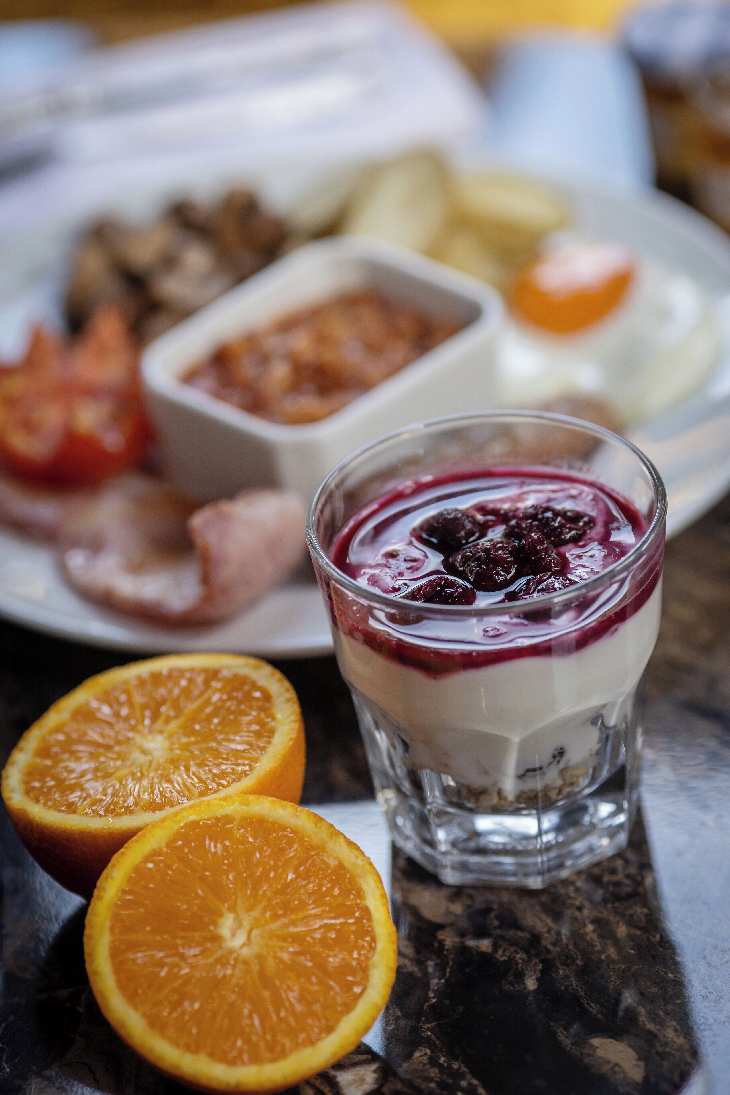 Et glas yoghurt med bær, en tallerken mad med kød og grøntsager, og to appelsinhælder på et bord.