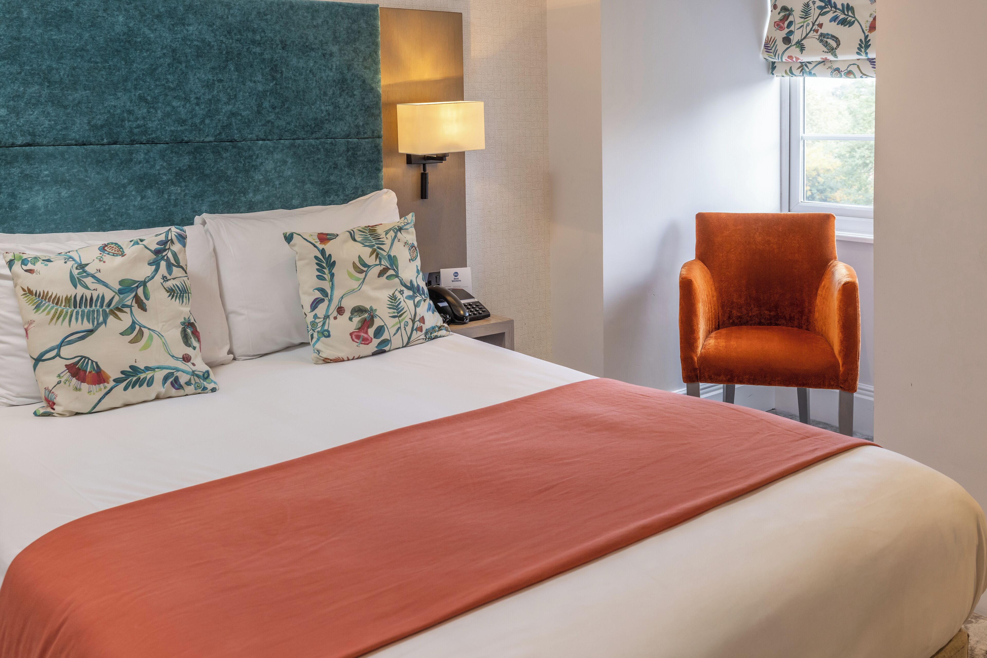 A neatly made bed with floral pillows, a teal headboard, a red throw blanket, a nightstand with a lamp, and a chair by the window.