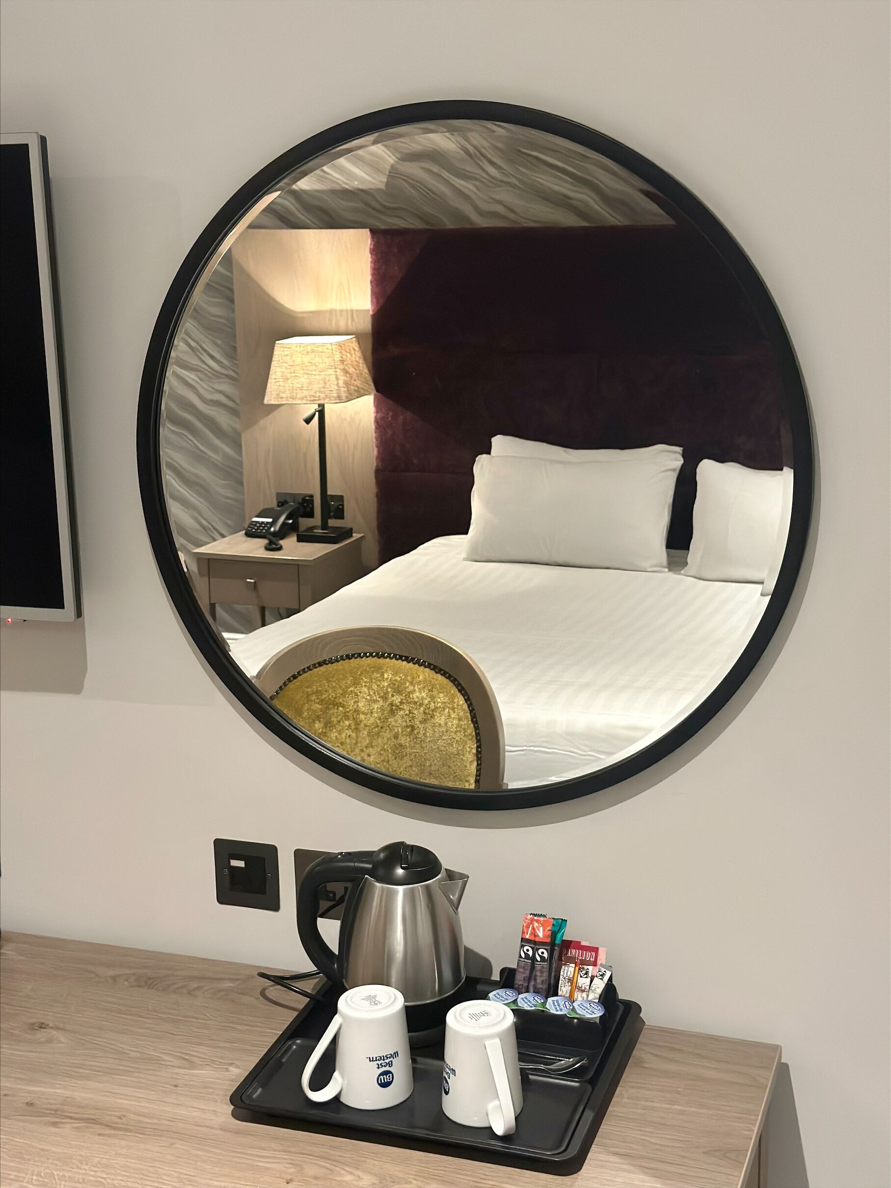 A hotel room with a large round mirror reflecting a bed, nightstand, lamp, and a coffee station with mugs and tea packets.