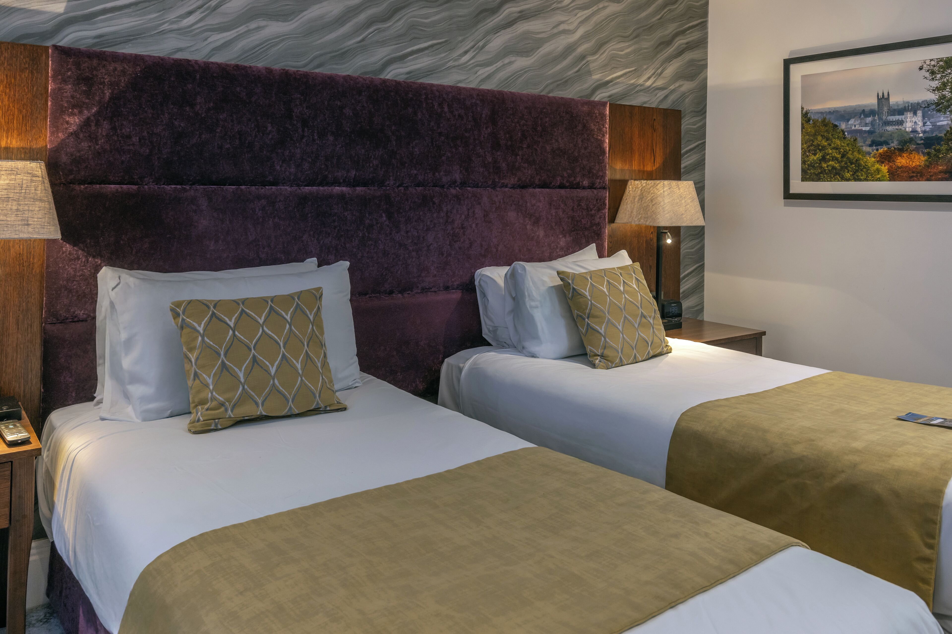 A hotel room with two beds, a purple headboard, and a cityscape painting on the wall.