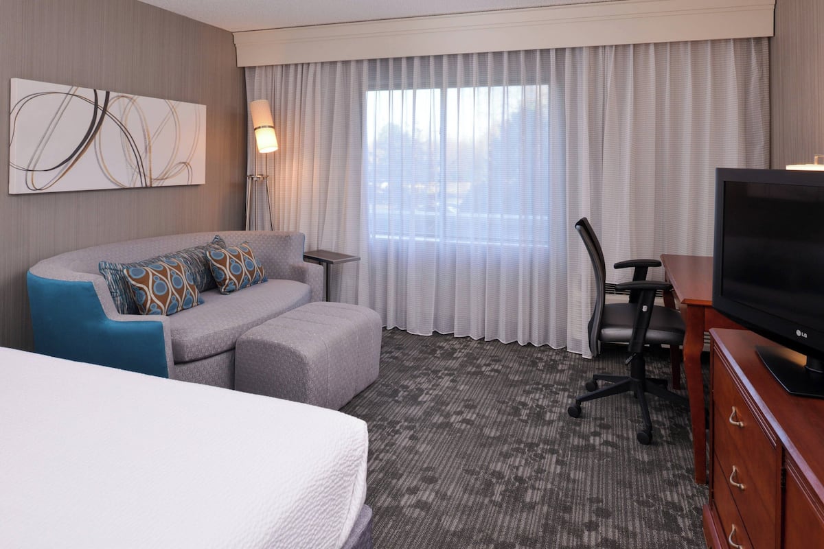 A hotel room with a bed, a grey sofa with patterned cushions, a desk with a chair, a television, and a large window with curtains.