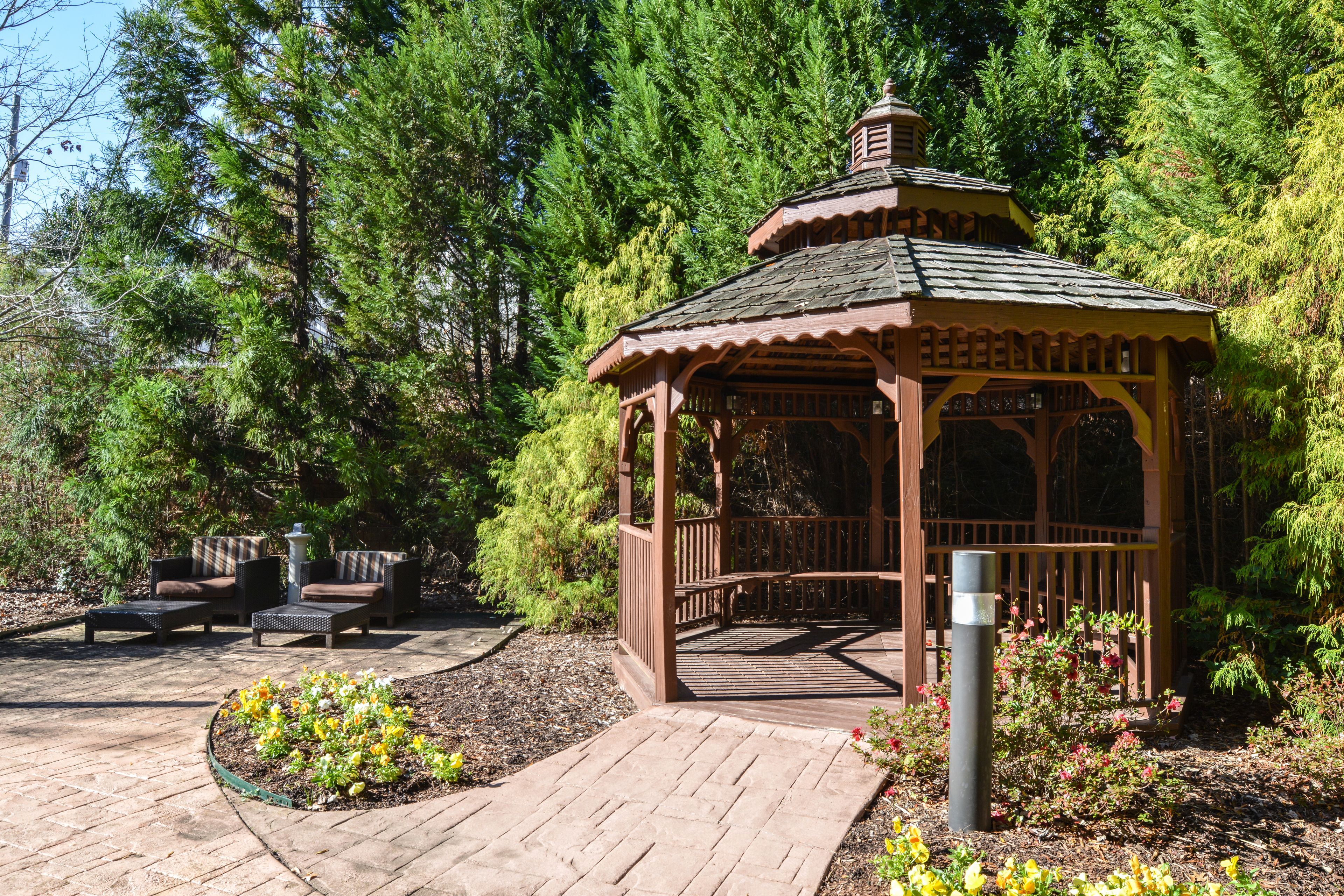 A wooden gazebo with a tiled roof, surrounded by a garden with flowers and a paved walkway.
