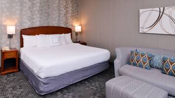 Habitación de hotel con una cama grande, dos mesitas de noche, un revistero, un sofá gris y una pared con papel tapiz estampado y una obra de arte abstracta.