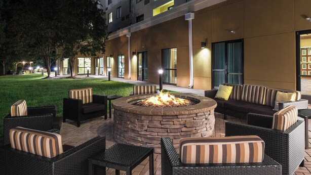 An outdoor seating area with wicker furniture, a stone fire pit, and a building with large windows in the background.