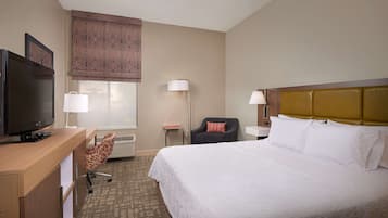 A hotel room with a bed, a desk with a television, a chair, a lamp, and a window with patterned curtains.