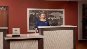 A reception desk with a framed picture of a mountainous landscape and the word "explore." in the background. - Hampton Inn Phoenix/Anthem (Phoenix)