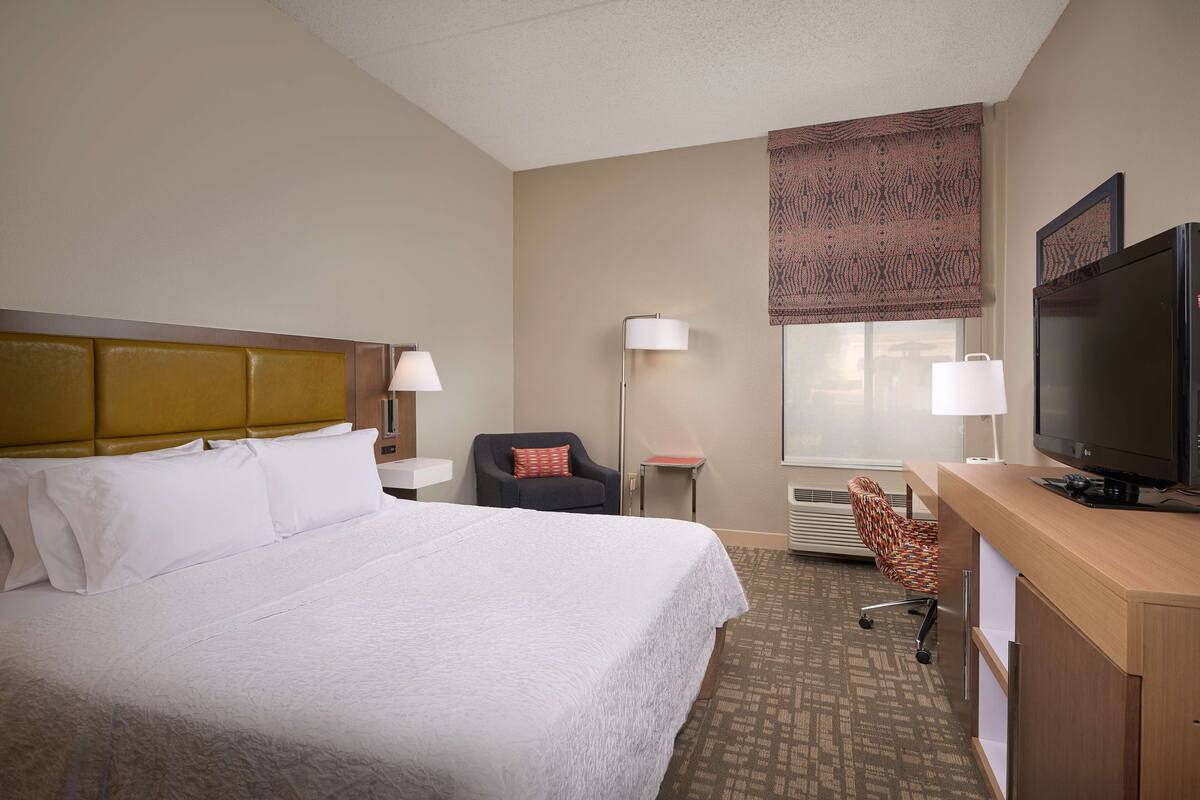 A hotel room with a large bed, a desk with a television, a chair, and a window with patterned curtains.
