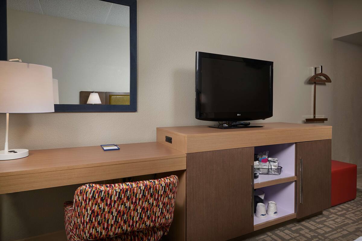 A hotel room with a wooden desk, a television, a lamp, a patterned chair, and a red ottoman.