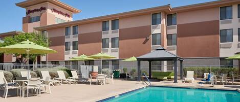 A hotel pool area with lounge chairs and umbrellas, a covered seating area, and a clear blue pool.