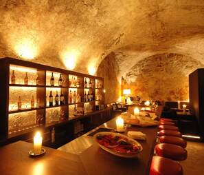 A bar with shelves of bottles and glasses, a candle-lit table with a plate of food, and red bar stools. - Hotel Misani (Celerina-Schlarigna)