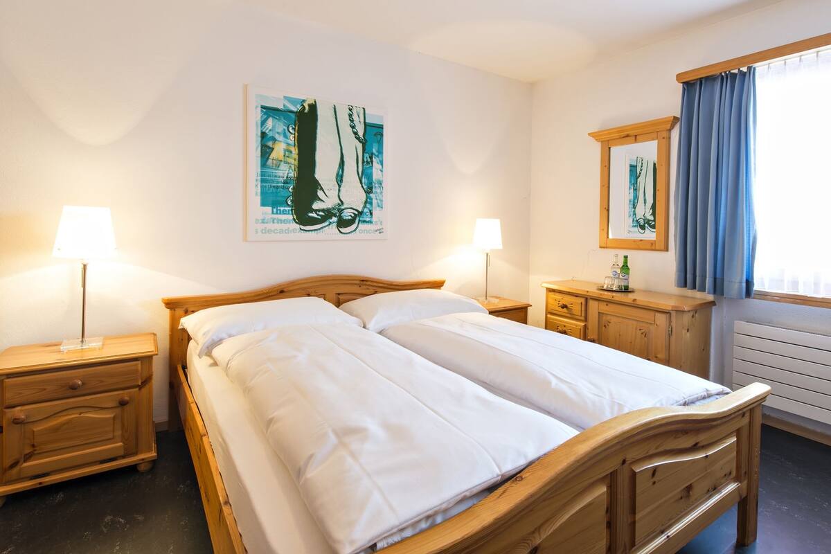 A hotel room with a wooden bed, two bedside tables with lamps, a dresser, and a window with curtains.