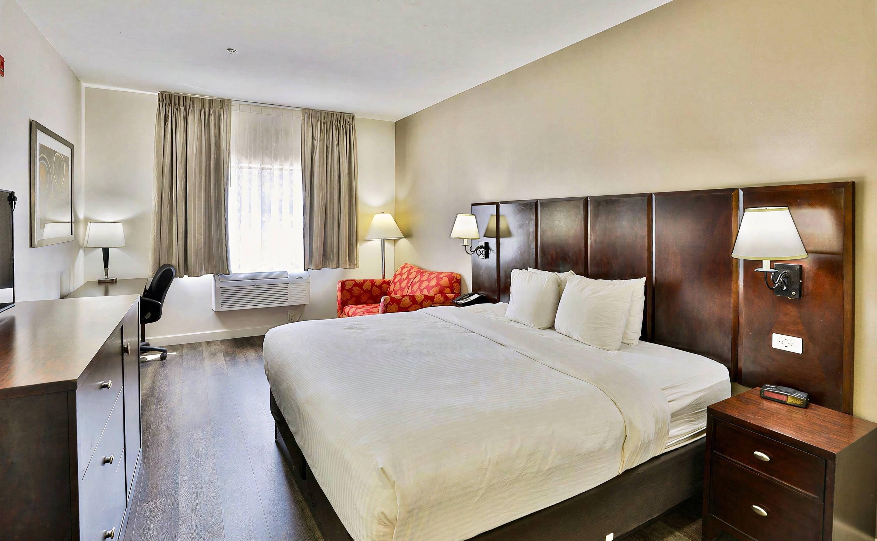 A hotel room with a large bed, bedside lamps, a desk with a chair, and a window with curtains.