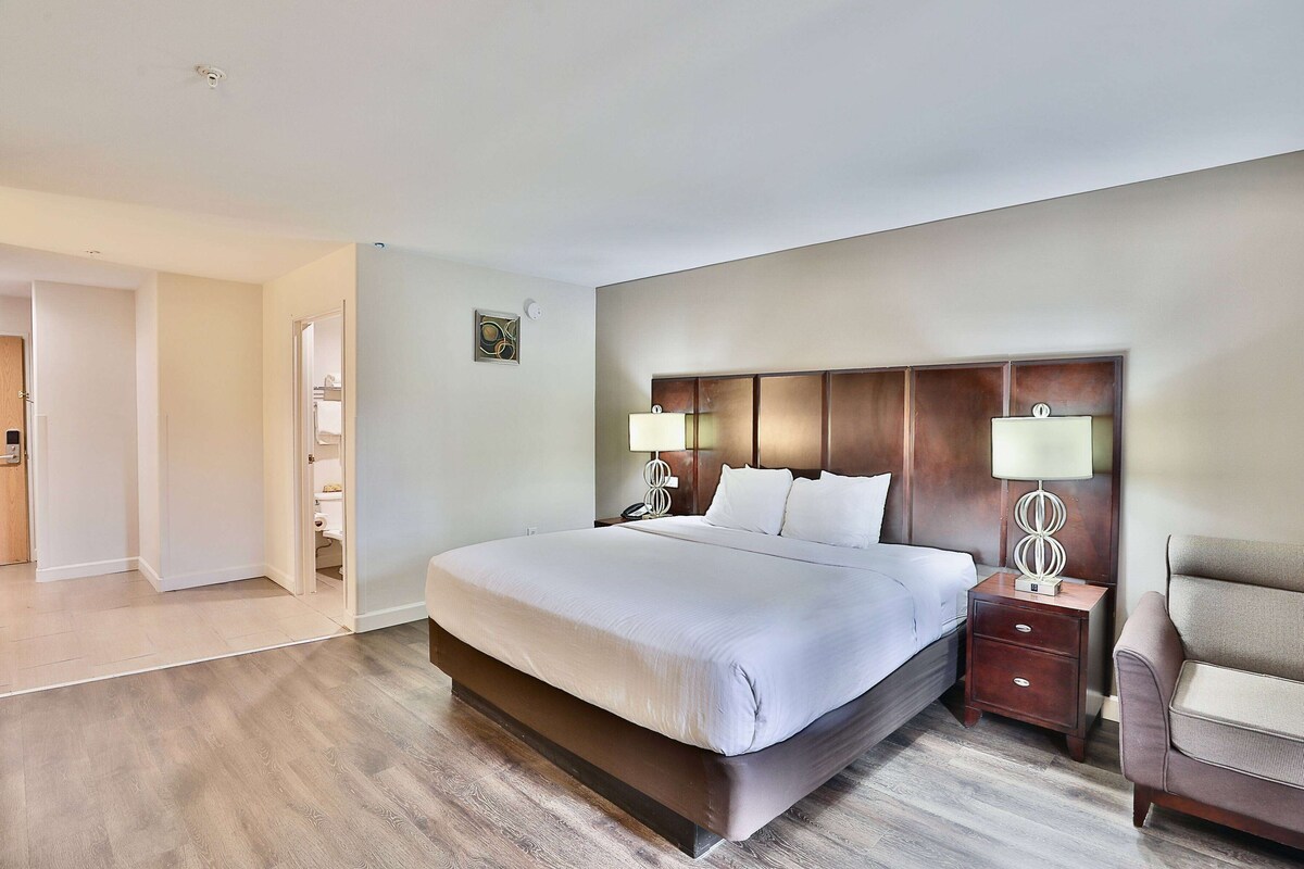 Una habitación de hotel moderna con una cama grande, cabecera de madera, dos mesitas de noche con lámparas, una silla y una puerta que da a otra habitación.