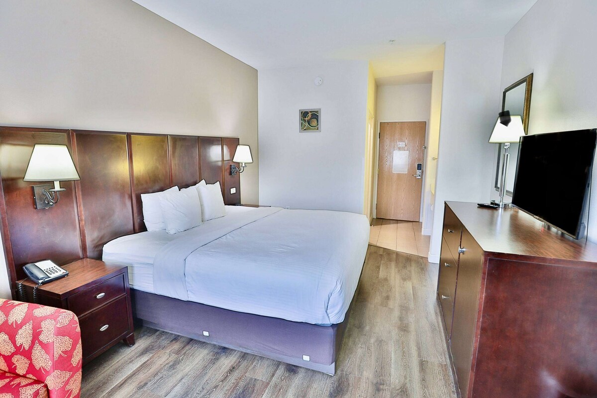 A hotel room with a large bed, bedside lamps, a TV, a wooden nightstand, and a red chair.