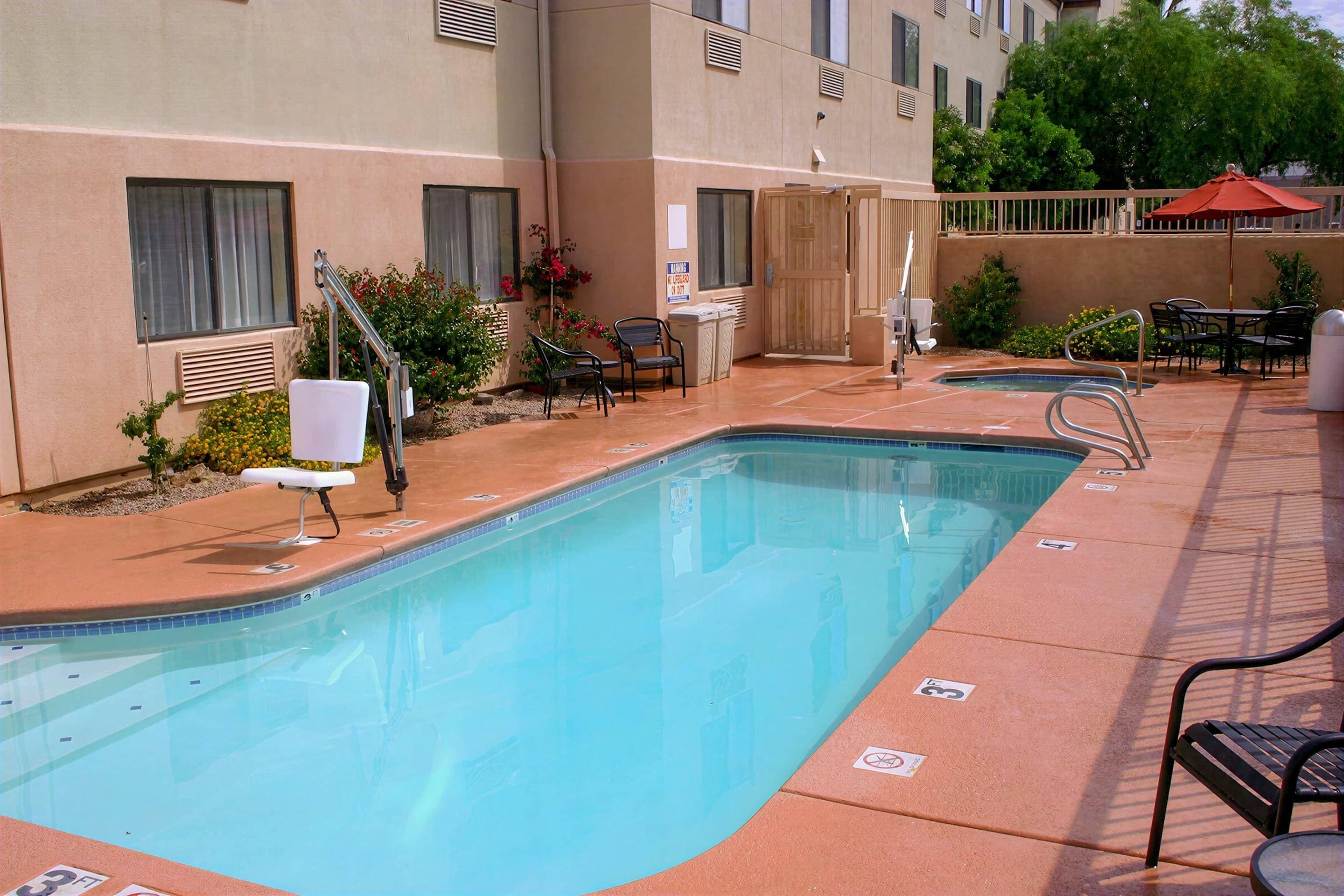 Una piscina con sillones de descanso y una mesa, rodeada por un edificio con ventanas y una puerta.