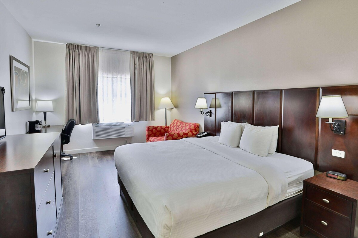 A hotel room with a large bed, a desk, a chair, a red sofa, two wall lamps, and a window with curtains.