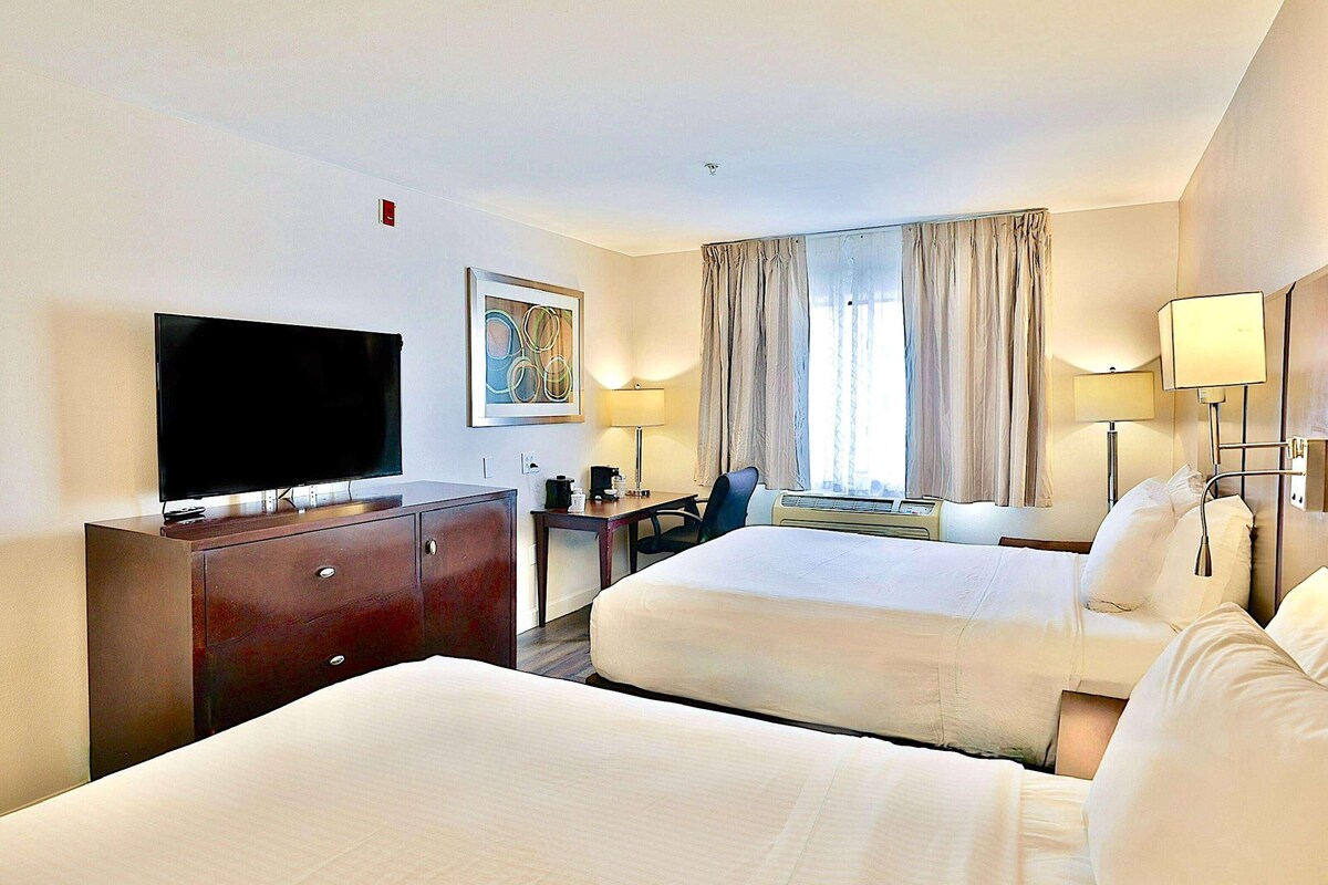 A hotel room with two beds, a desk, a TV, and a window with curtains.