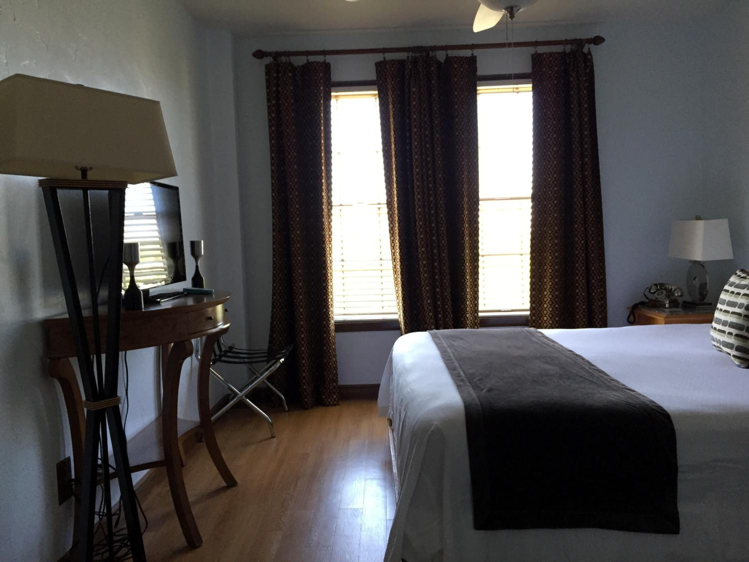 A hotel room with a bed, a desk with a computer, a chair, a lamp, and a window with curtains.