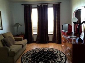 Una sala de estar con un sofá, un televisor sobre un mueble de madera, un tapete estampado y cortinas.