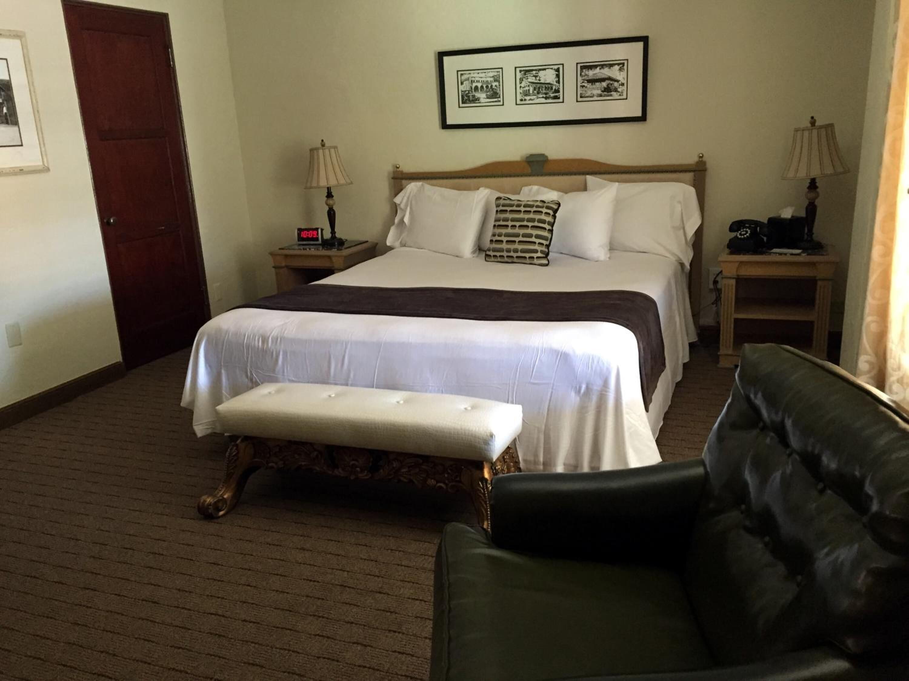 A hotel room with a large bed, a bench, a black leather chair, two bedside tables with lamps, and a framed picture above the bed.