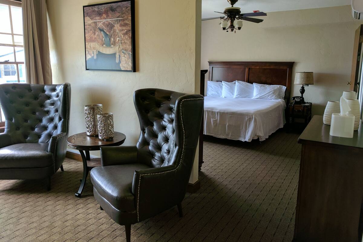 A hotel room with a bed, two leather armchairs, a small table with decorative items, a ceiling fan, and a painting on the wall.