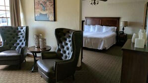 A hotel room with a bed, two leather armchairs, a small table with decorative items, a ceiling fan, and a painting on the wall. - Boulder Dam Hotel (Boulder City)