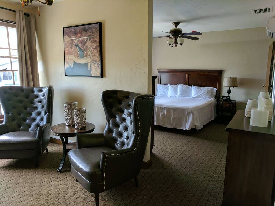 A hotel room with a bed, two leather armchairs, a small table with decorative items, a ceiling fan, and a painting on the wall.