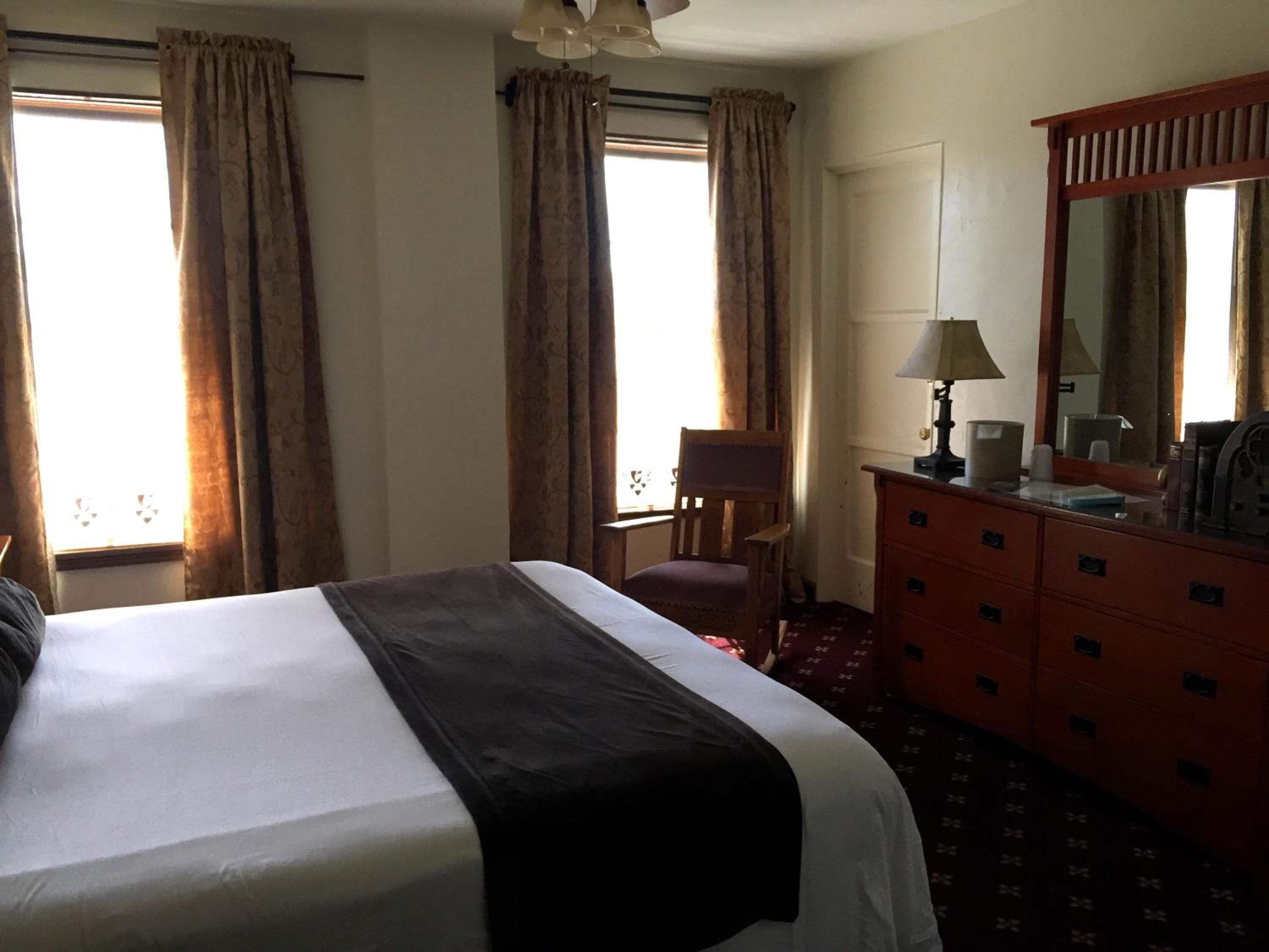 A hotel room with a large bed, a wooden chair, a dresser with drawers, a mirror, and two windows with curtains.