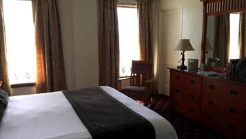 A hotel room with a large bed, a wooden chair, a dresser with drawers, a mirror, and two windows with curtains.
