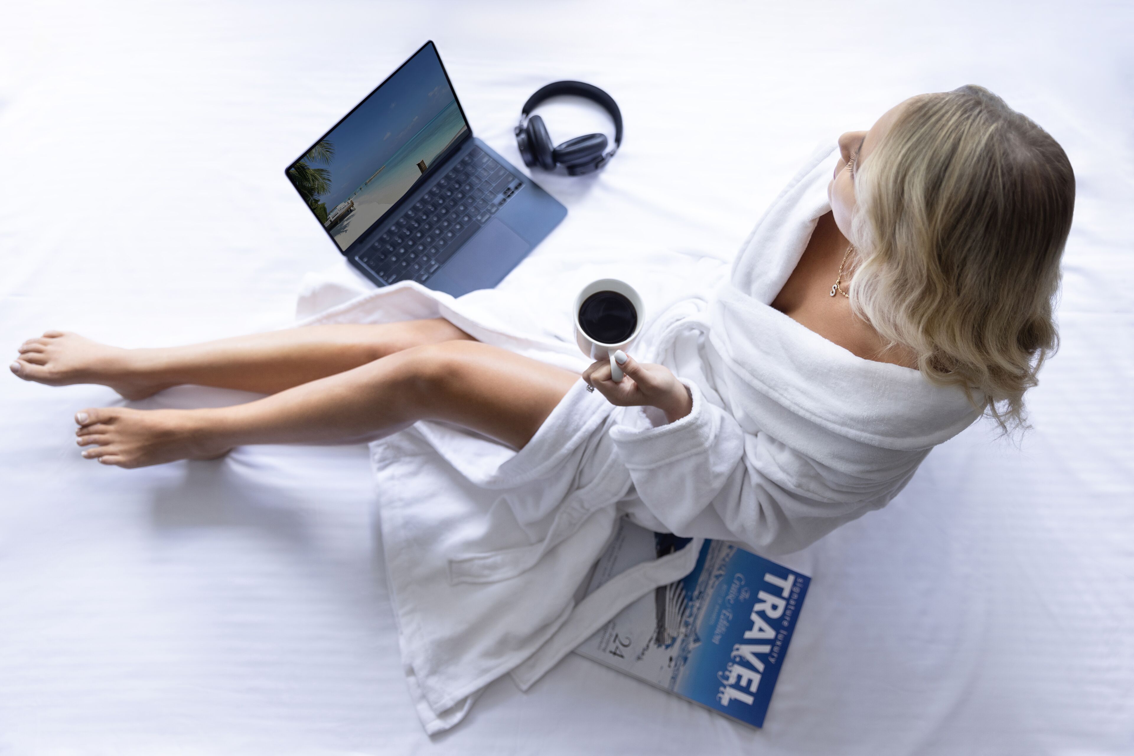 A person sitting on a bed with a laptop, headphones, and a travel book.