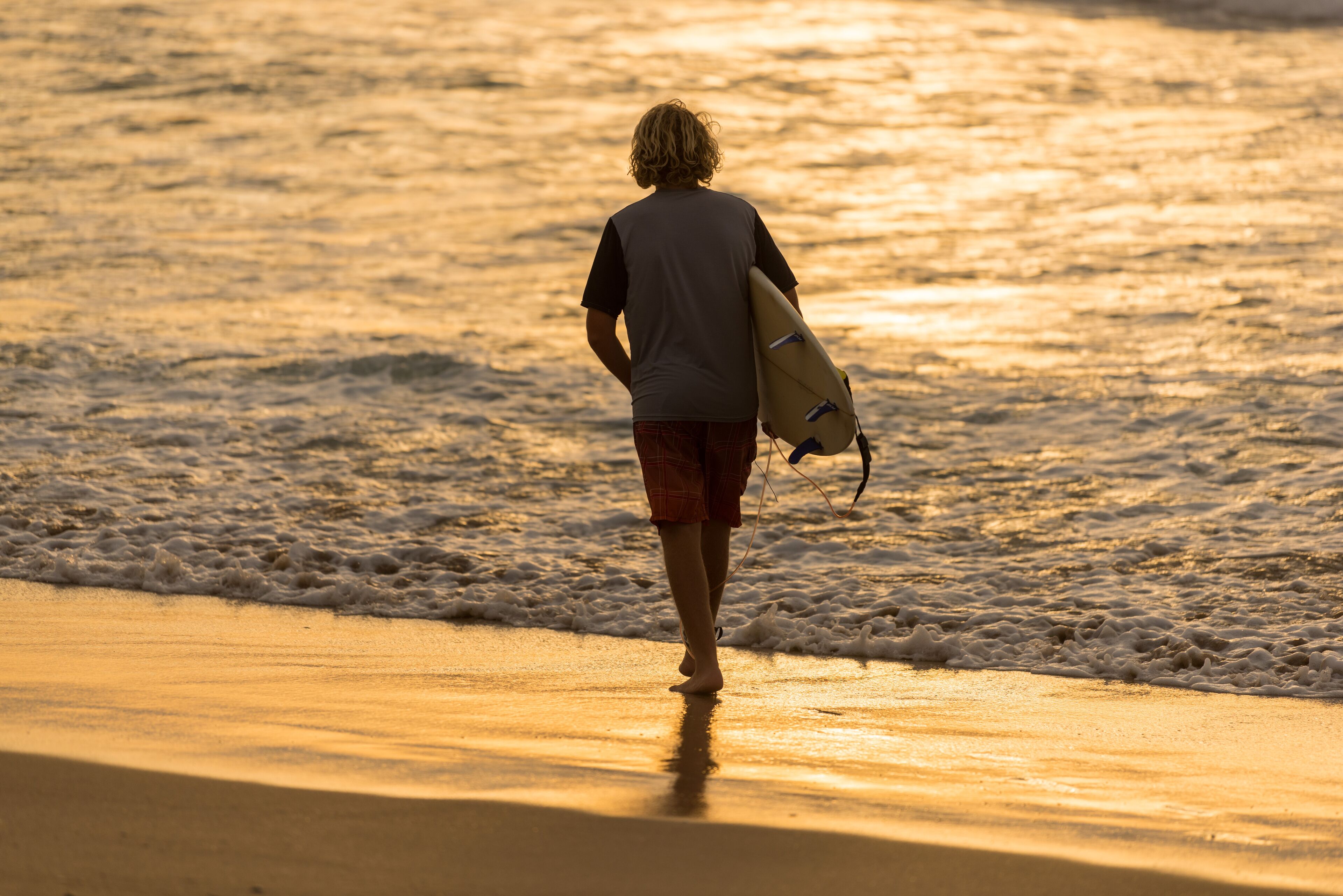 Une personne se promène sur une plage, une planche de surf à la main, vêtue d’un t-shirt gris et d’un short rouge.