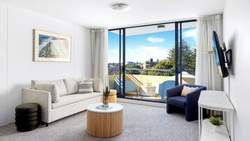 A modern living room with a sofa, a round coffee table, and a blue armchair. There is a large window with a view of the city.