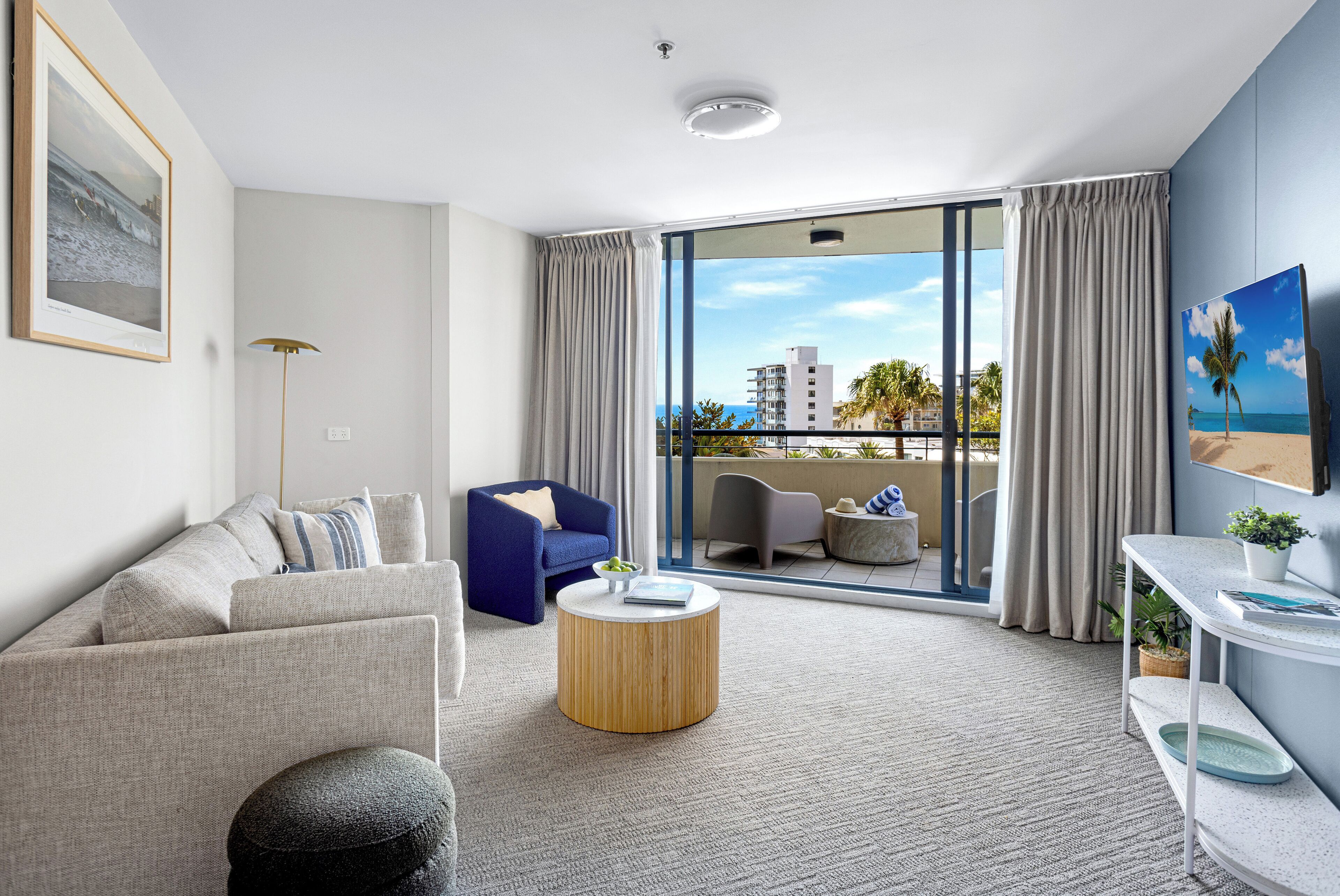 A modern living room with a sofa, a blue armchair, a round coffee table, and a view of the balcony with a cityscape and palm trees.