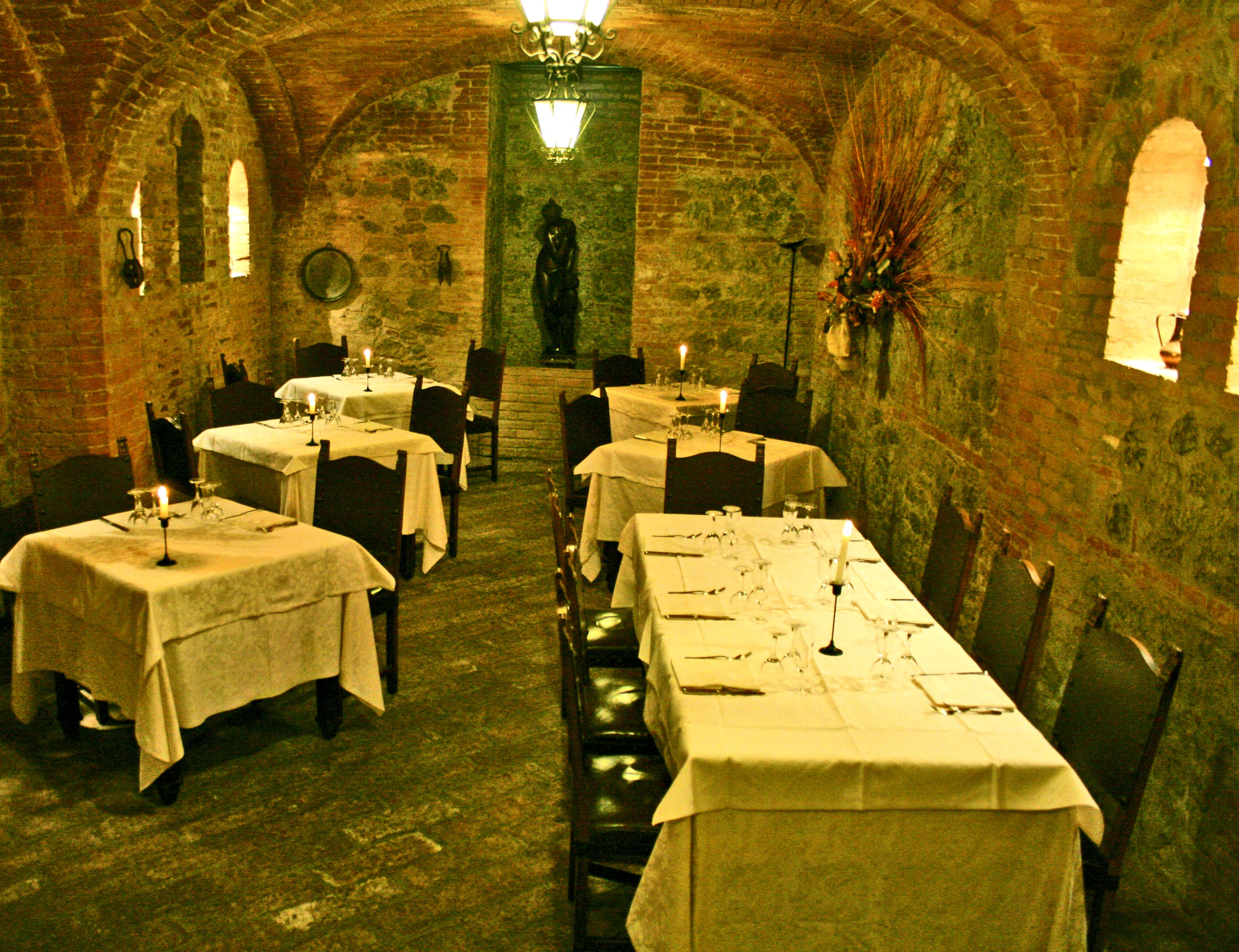 A dimly lit, rustic dining area with brick walls and arched ceilings, featuring tables set for dining with candles and wine glasses.