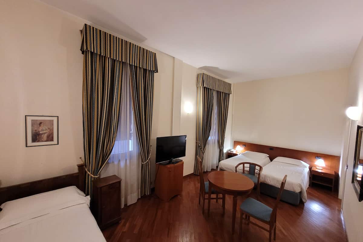 A hotel room with a wooden floor, a TV, a desk, two beds, a chair, and curtains.
