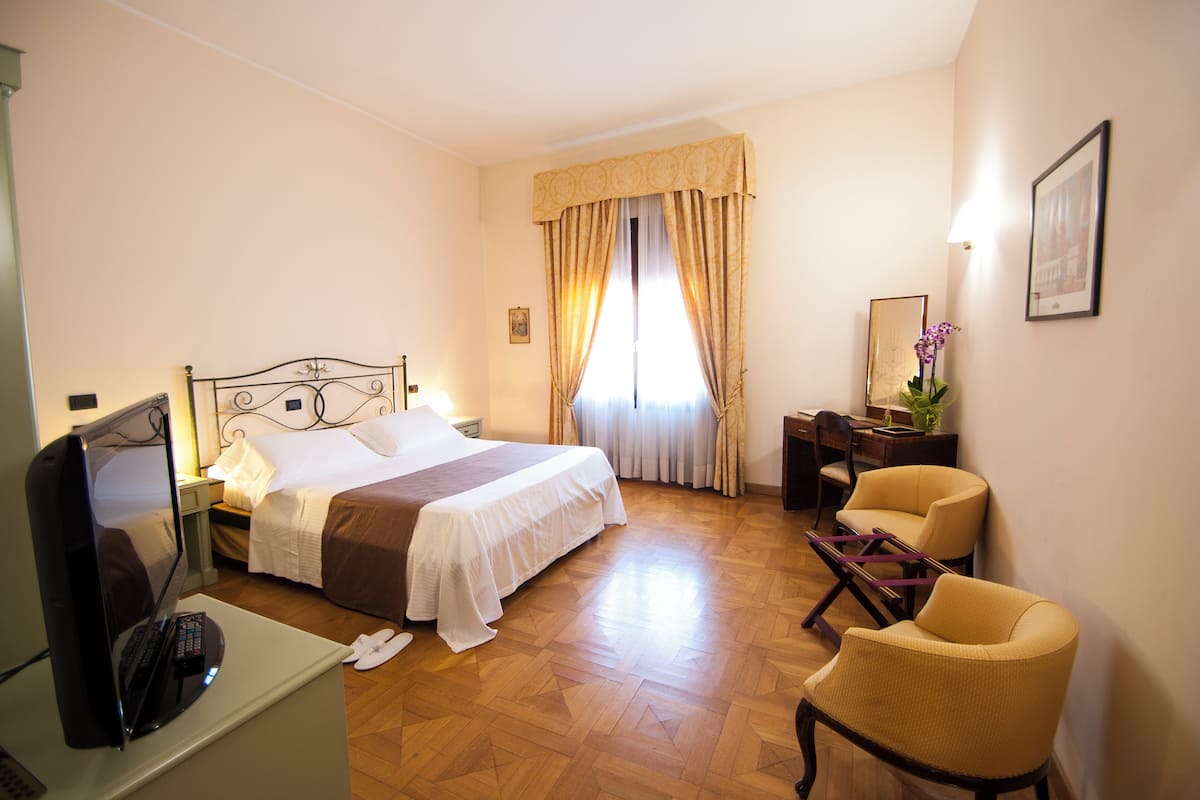 A hotel room with a bed, a desk with a computer, a chair, and a window with curtains.