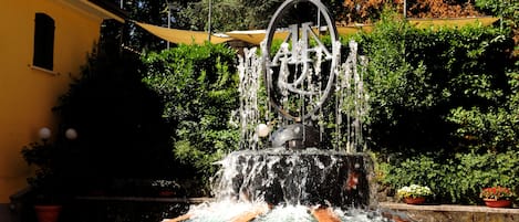 A fountain with water cascading into a pool, surrounded by greenery and a yellow building.