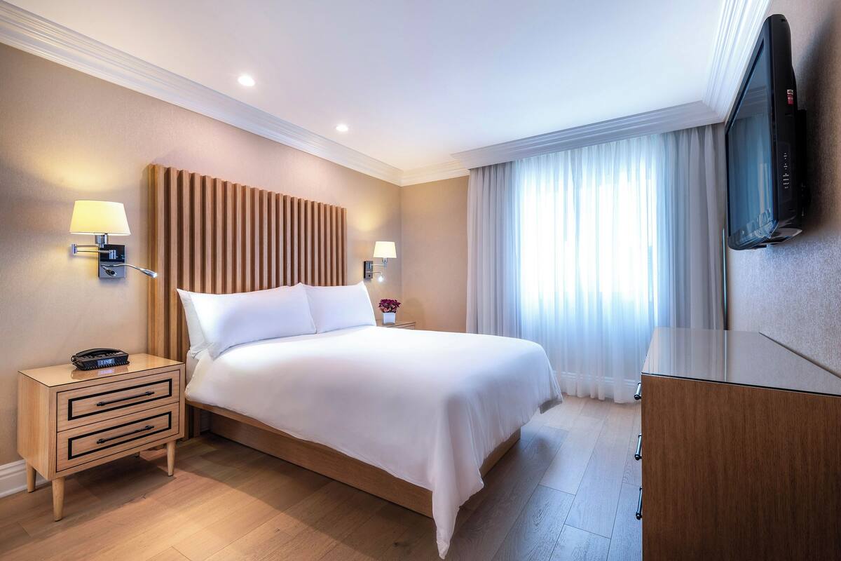 A hotel room with a large bed, bedside lamps, a flat-screen TV, and a wooden dresser.