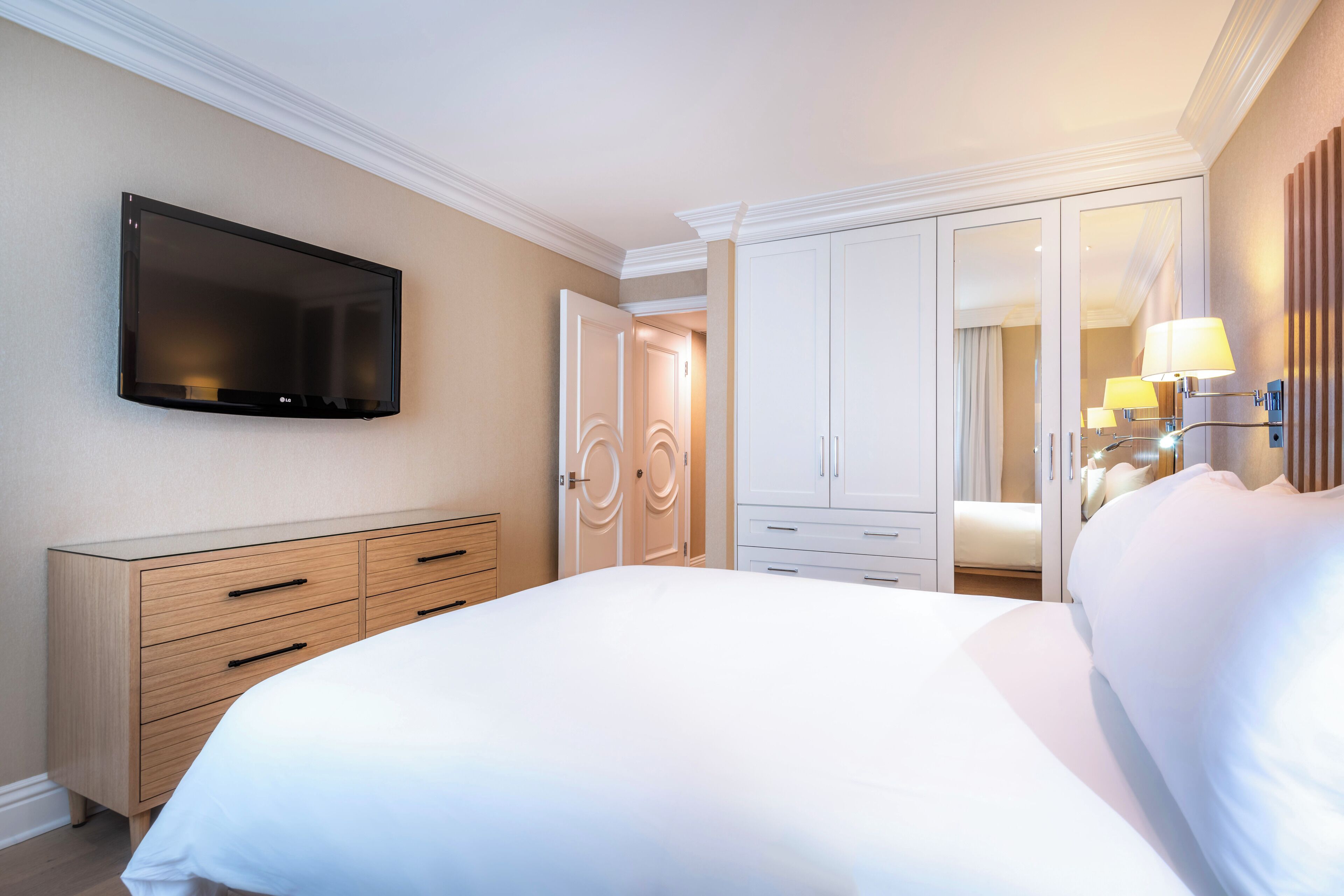 A hotel room with a large bed, a TV mounted on the wall, a wardrobe, and a mirror with lights.