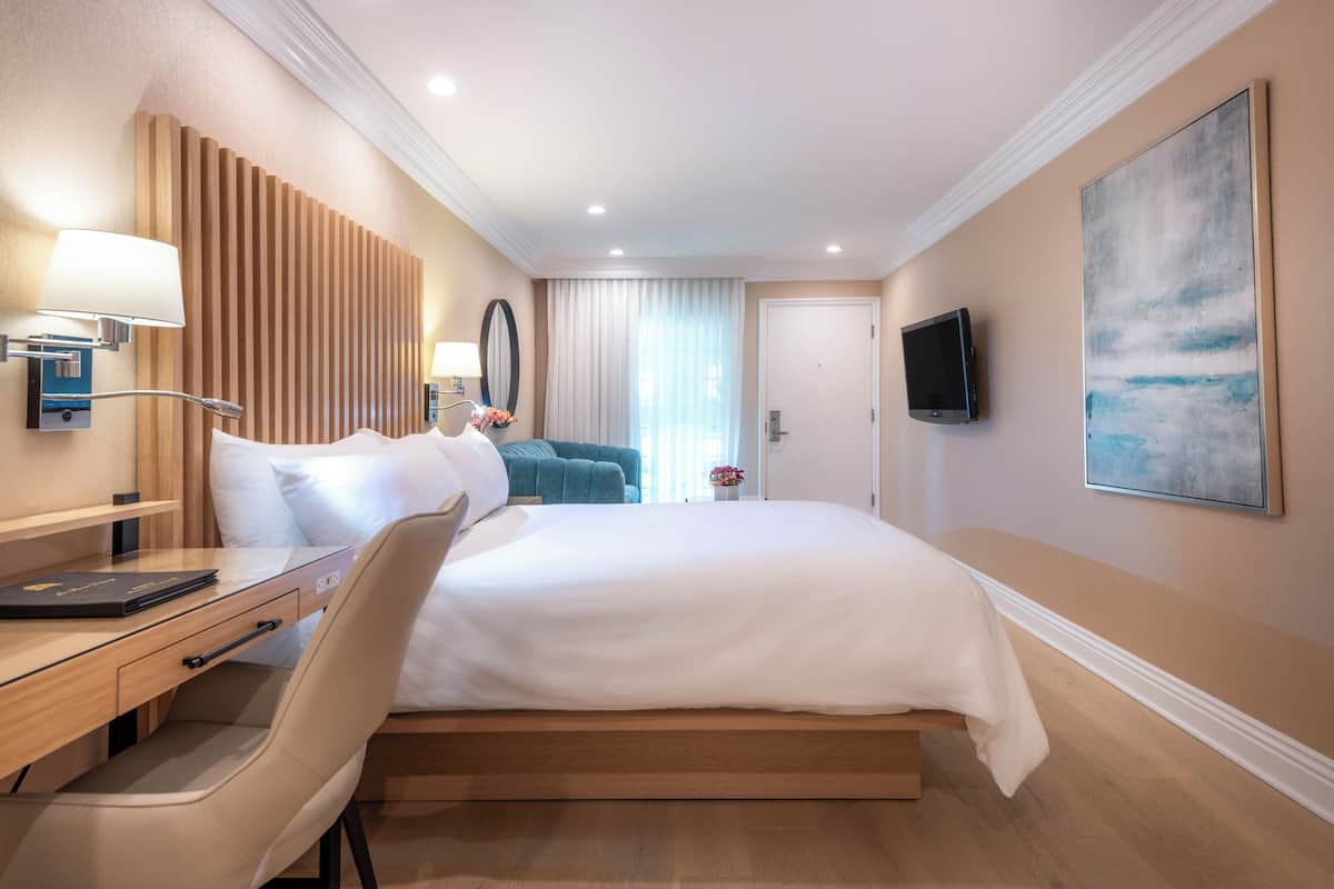 A hotel room with a bed, desk, chair, lamp, TV, and artwork.