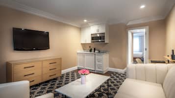 Een moderne hotelkamer met een flatscreen televisie, een eettafel met een bloemenvaas, een bank, een keuken met witte kasten en een magnetron, en een kamerbreed tapijt met dessin.