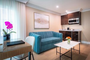 Una sala moderna con un sofá azul, una mesa de centro de vidrio, una mesa de comedor con un libro y una cocina con armarios de madera oscura y un microondas.