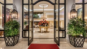 Une entrée somptueuse, avec un tapis rouge menant à un hall d’accueil où trône une grande table dorée agrémentée de compositions florales, le tout entouré de meubles élégants et surmonté d’un lustre.