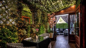 Un espace de restauration intérieur avec une table en verre, des chaises en osier et une vue sur un jardin luxuriant, visible à travers une porte ouverte.