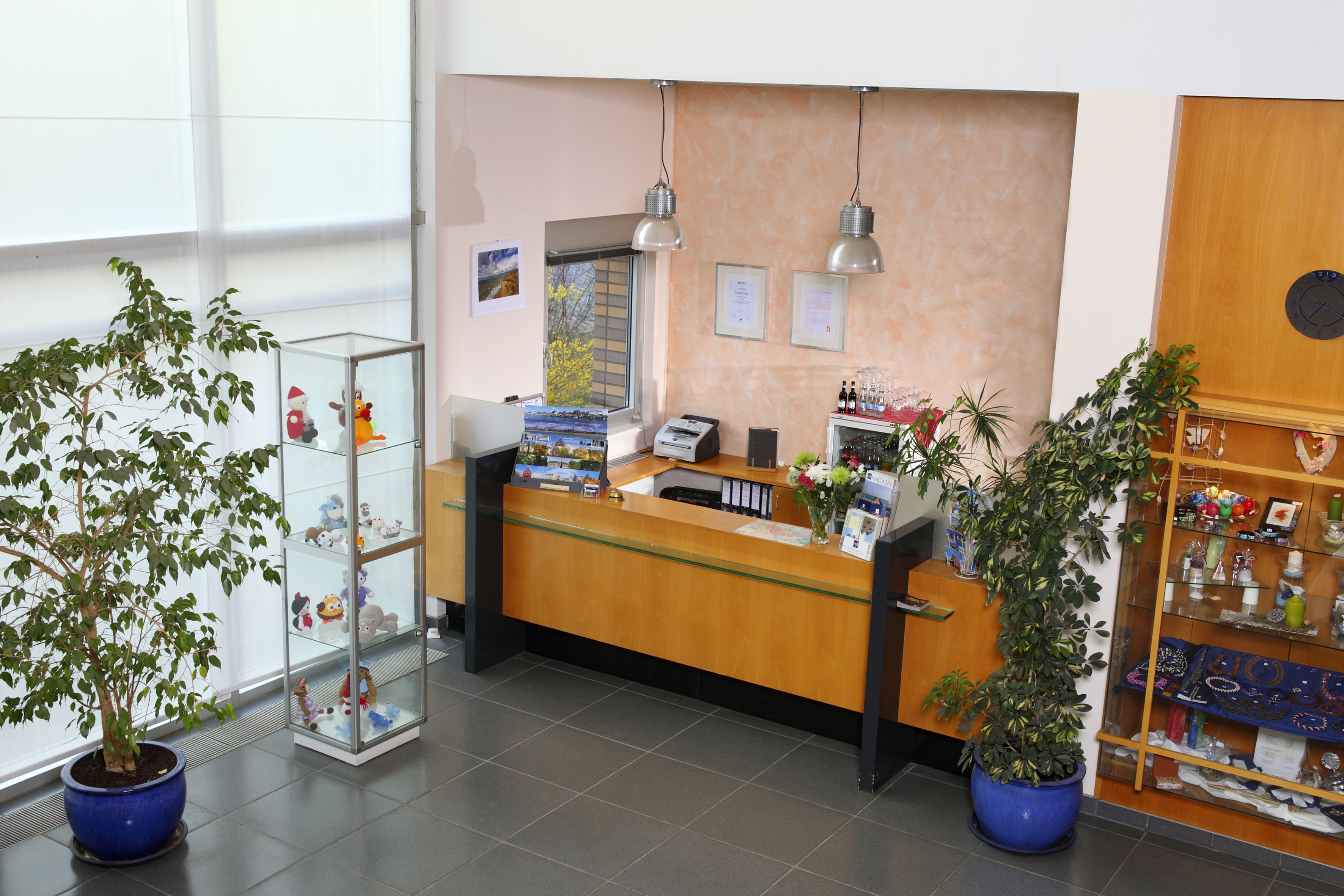 A reception desk with a computer, a printer, and a glass display case with items. There are two potted plants, a wooden cabinet with decorative items, and a wall clock.