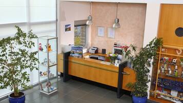 A reception desk with a computer, a printer, and a glass display case with items. There are two potted plants, a wooden cabinet with decorative items, and a wall clock.